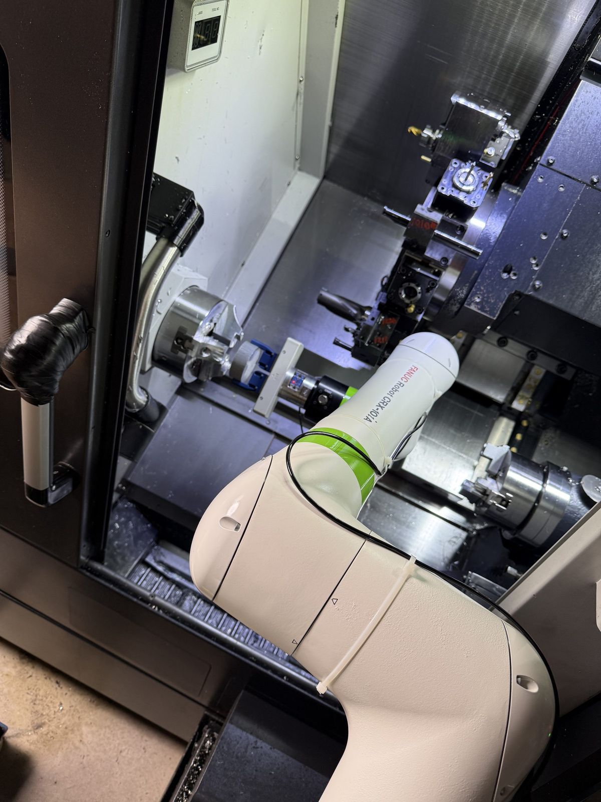 A FANUC CRX-10iA collaborative robot reaching in through the open door of a CNC lathe, with the machine’s turret, tooling and chuck in frame behind the arm.